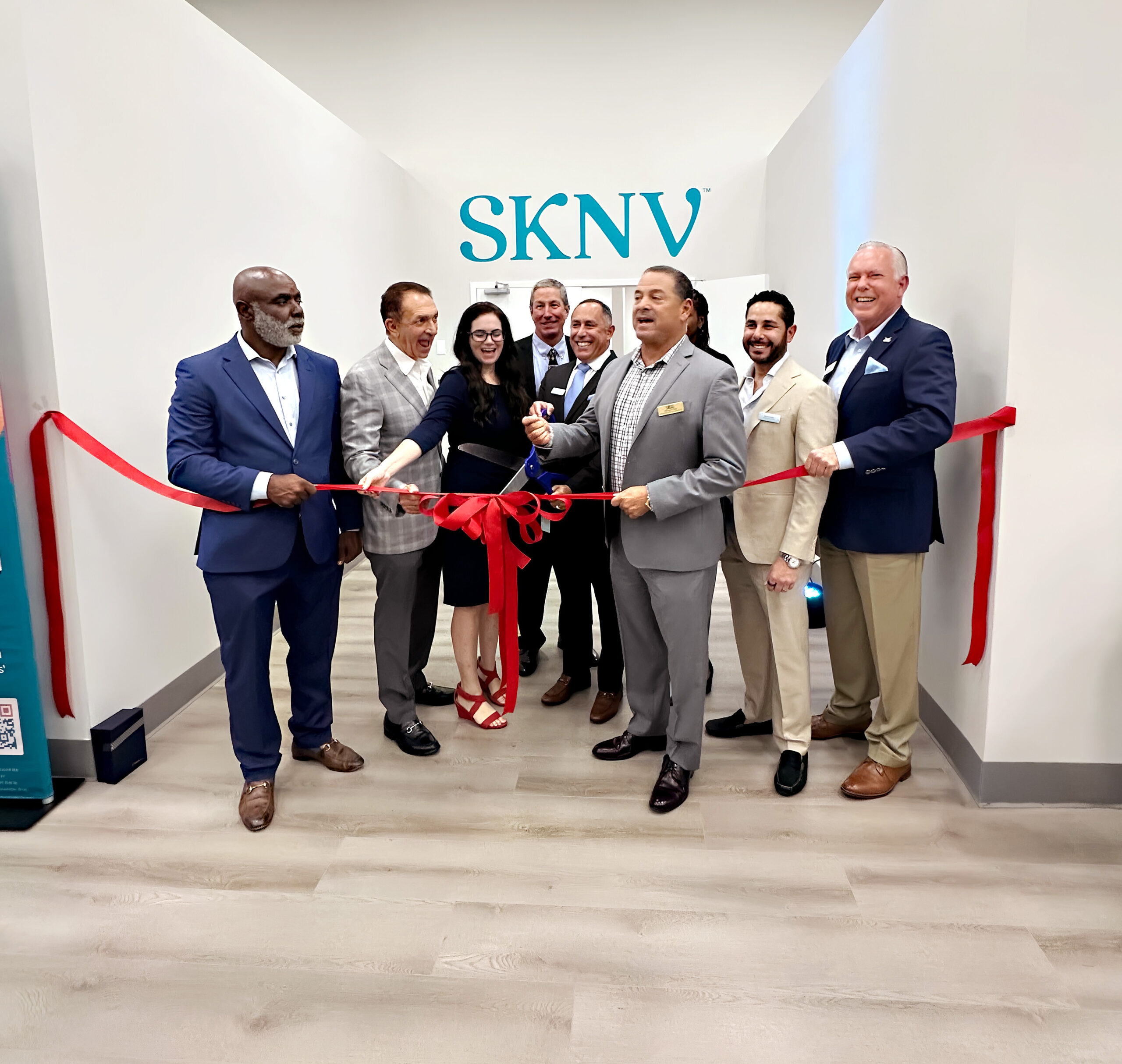 A group of eight people stand together holding a large red ribbon during a ribbon-cutting ceremony, with the SKNV logo displayed prominently in the background.